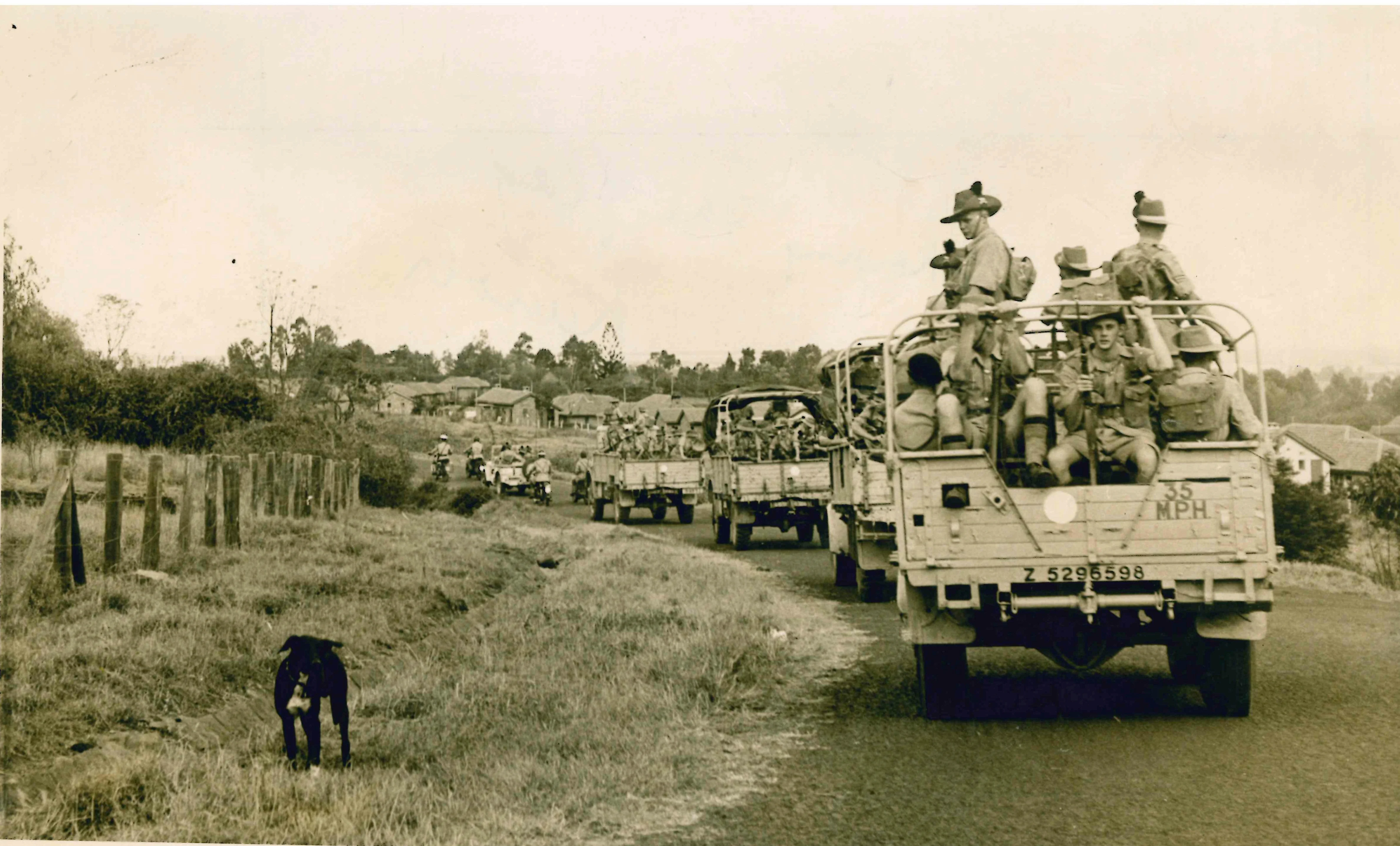 Historic sepia-toned photo showing a convoy of military trucks carrying armed British soldiers along a rural road through a Kenyan settlement. A dog stands beside the road.