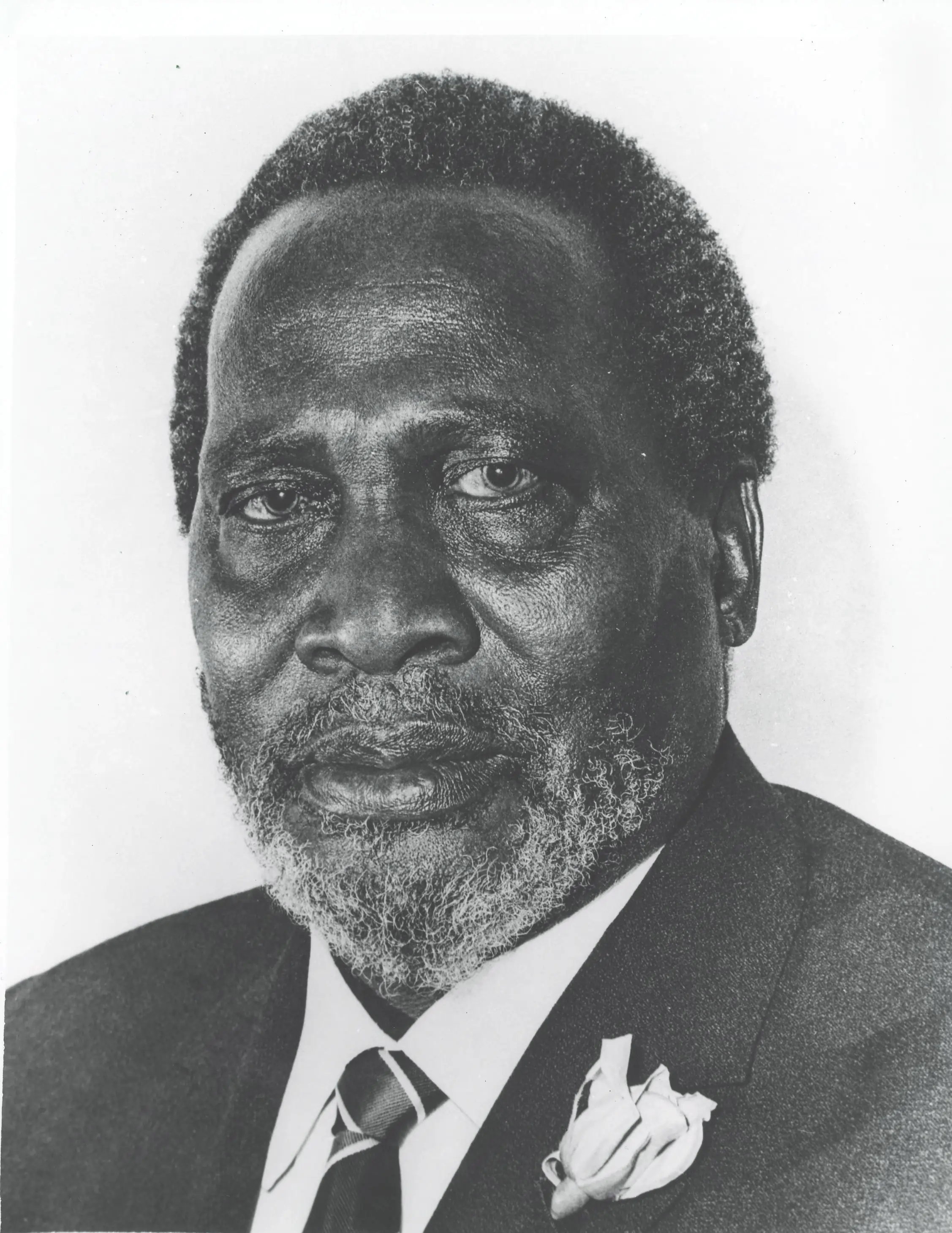 Black-and-white close-up portrait photo of President Jomo Kenyatta wearing a suit, striped tie, and lapel flower, looking directly at the camera.