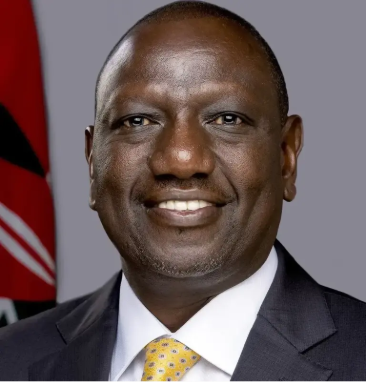 Portrait of a smiling President William Ruto in a dark suit, white shirt and yellow patterned tie, against a light gray background with part of the Kenyan flag visible on the left side.