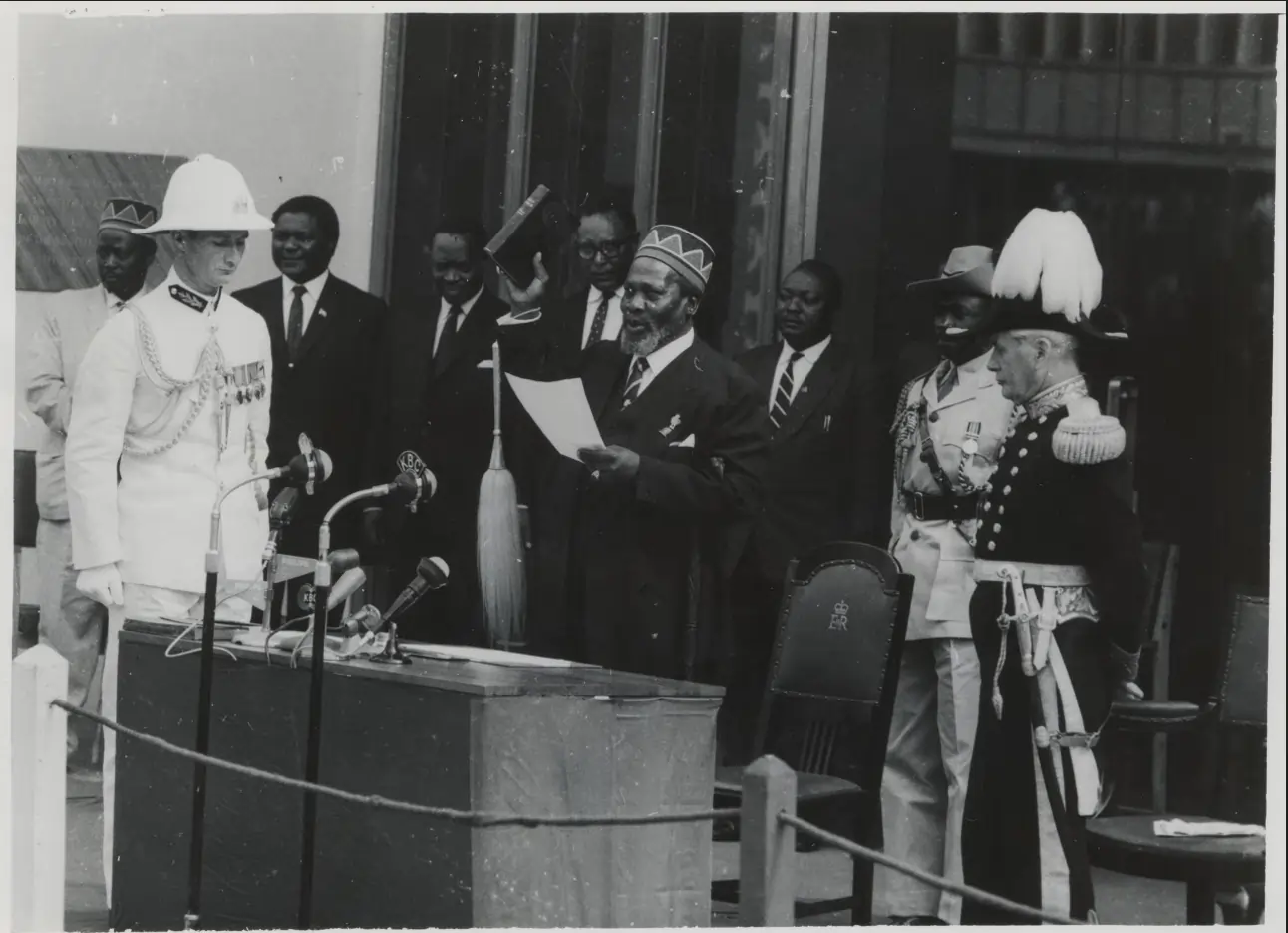 Historic black and white photo of Jomo Kenyatta lifting the bible and oath of office statement while surrounded by military officers, the ceremonial guard and government officials.