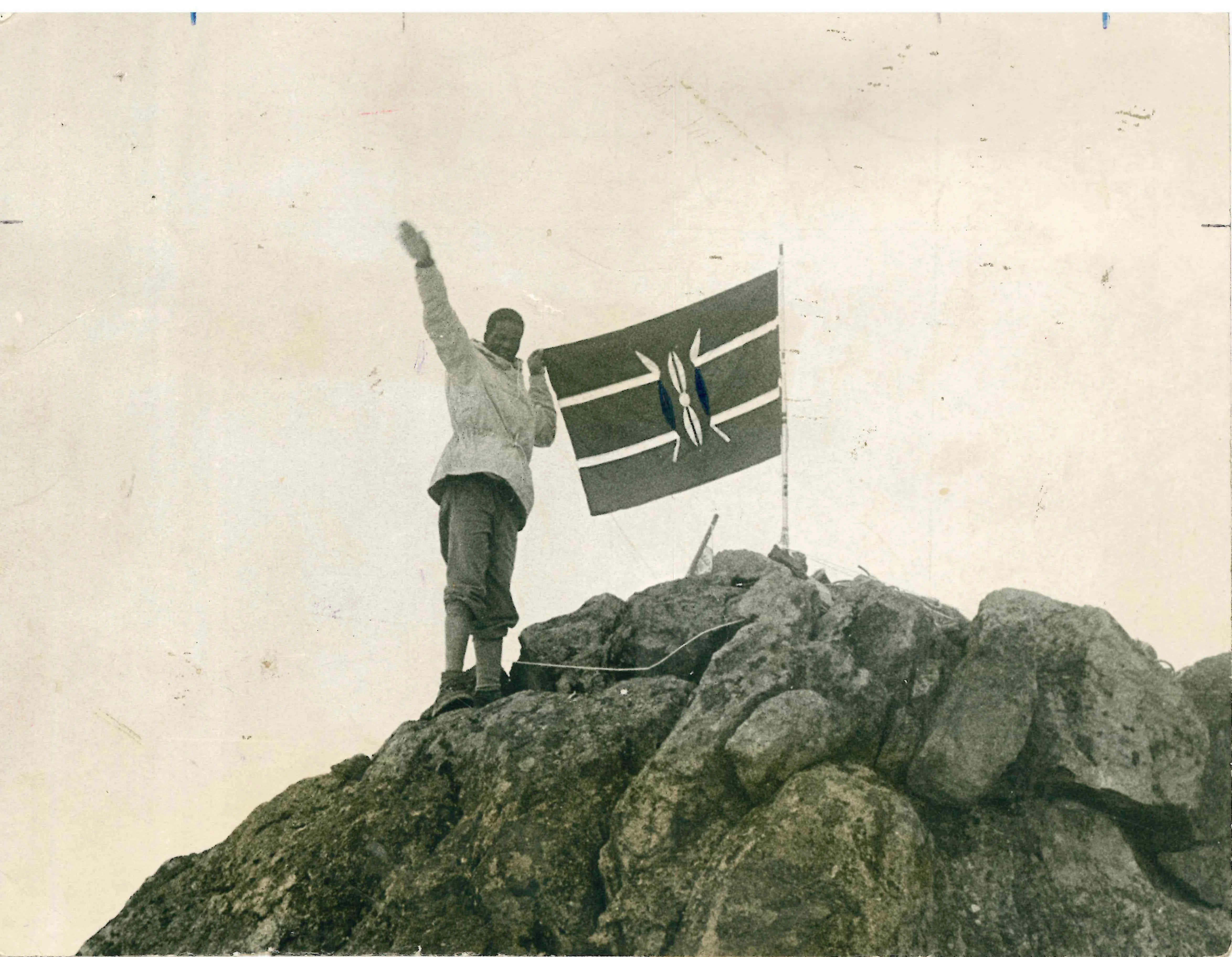 Black and white photo of Kisoi Munyao standing atop a rocky peak of Mt Kenya raising his arm beside an early national flag fluttering in the wind against an open sky.