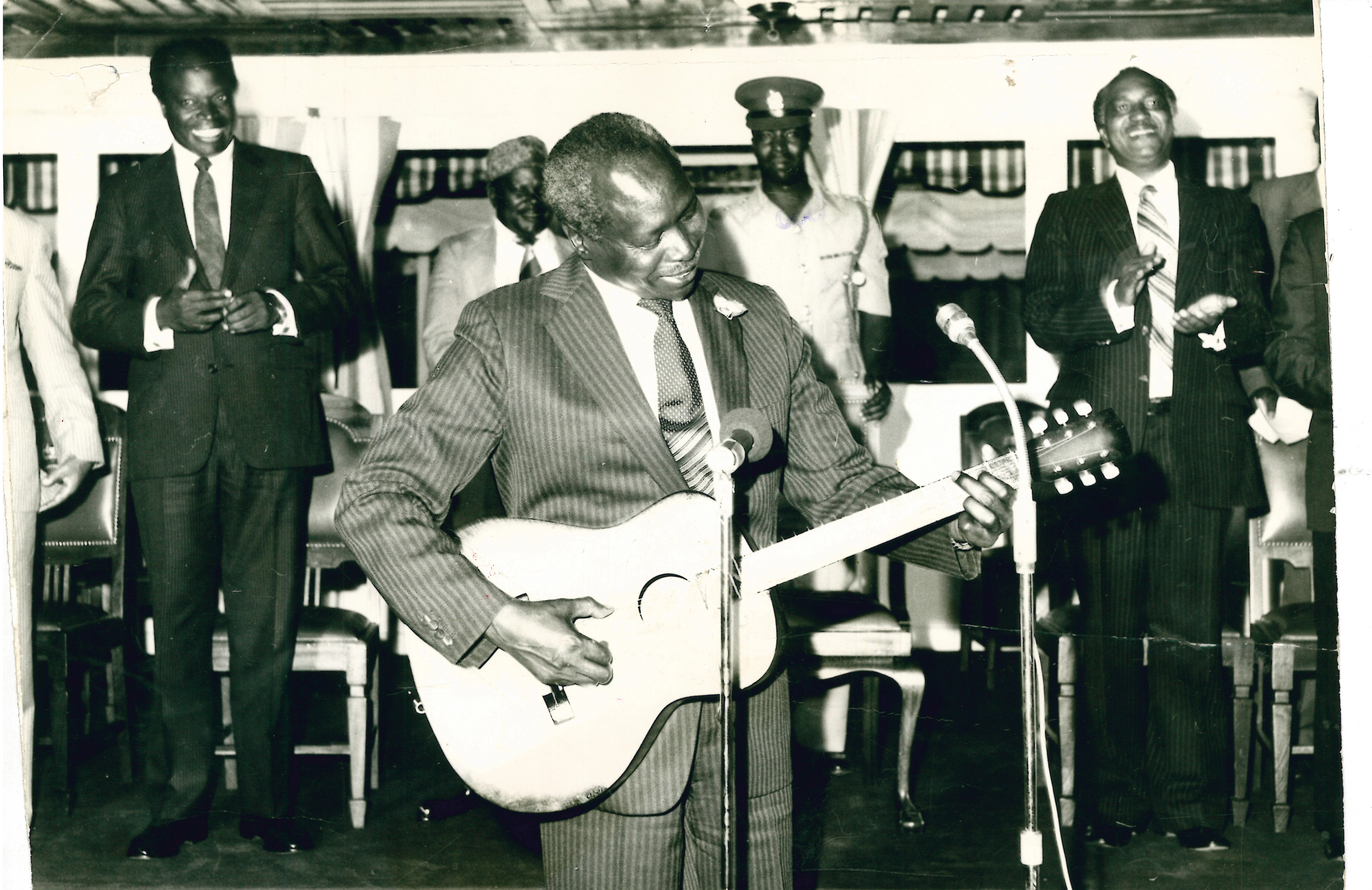 President Daniel Arap Moi tries his hand to play a guitar presented to him by Musician Joseph Kamaru as Minister for Culture Jeremiah Nyagah looks on