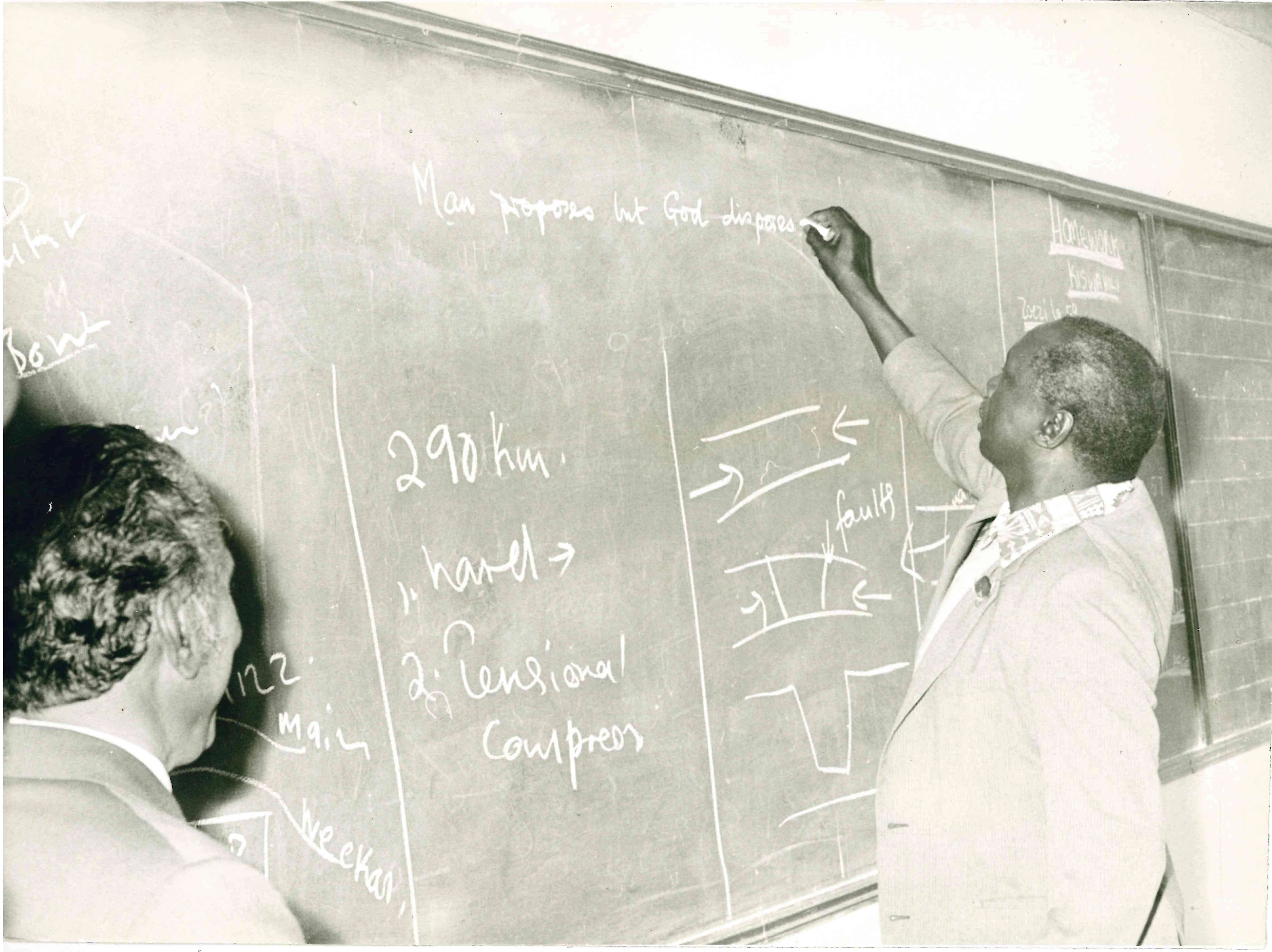President Daniel arap Moi wearing a light-coloured suit and patterned shirt, stretches his right hand up to write near the top of a large blackboard above other writings and sketches. Looking on from the foreground of his left hand side is a man in a suit.