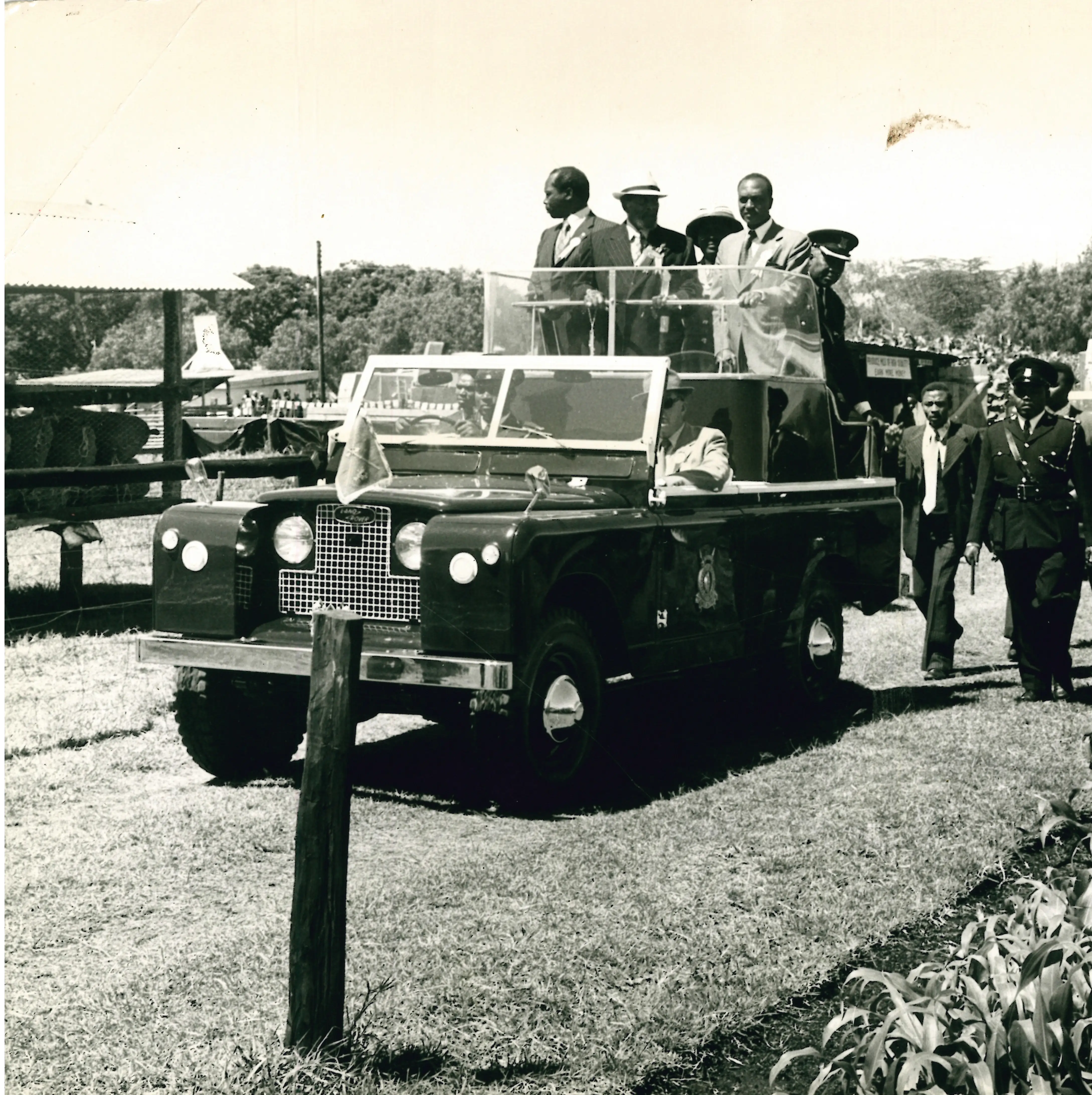 Historic black-and-white photo showing President Jomo Kenyatta standing at the centre of an open-top ceremonial Land Rover, flanked by Vice President Moi to his right, and the Agriculture minister Jeremiah Nyagah. They are surrounded by a battery of senior security officers and bodyguards. At the bottom right of the photo is a small section of a maize farm, given the event was an agricultural show in Nakuru.