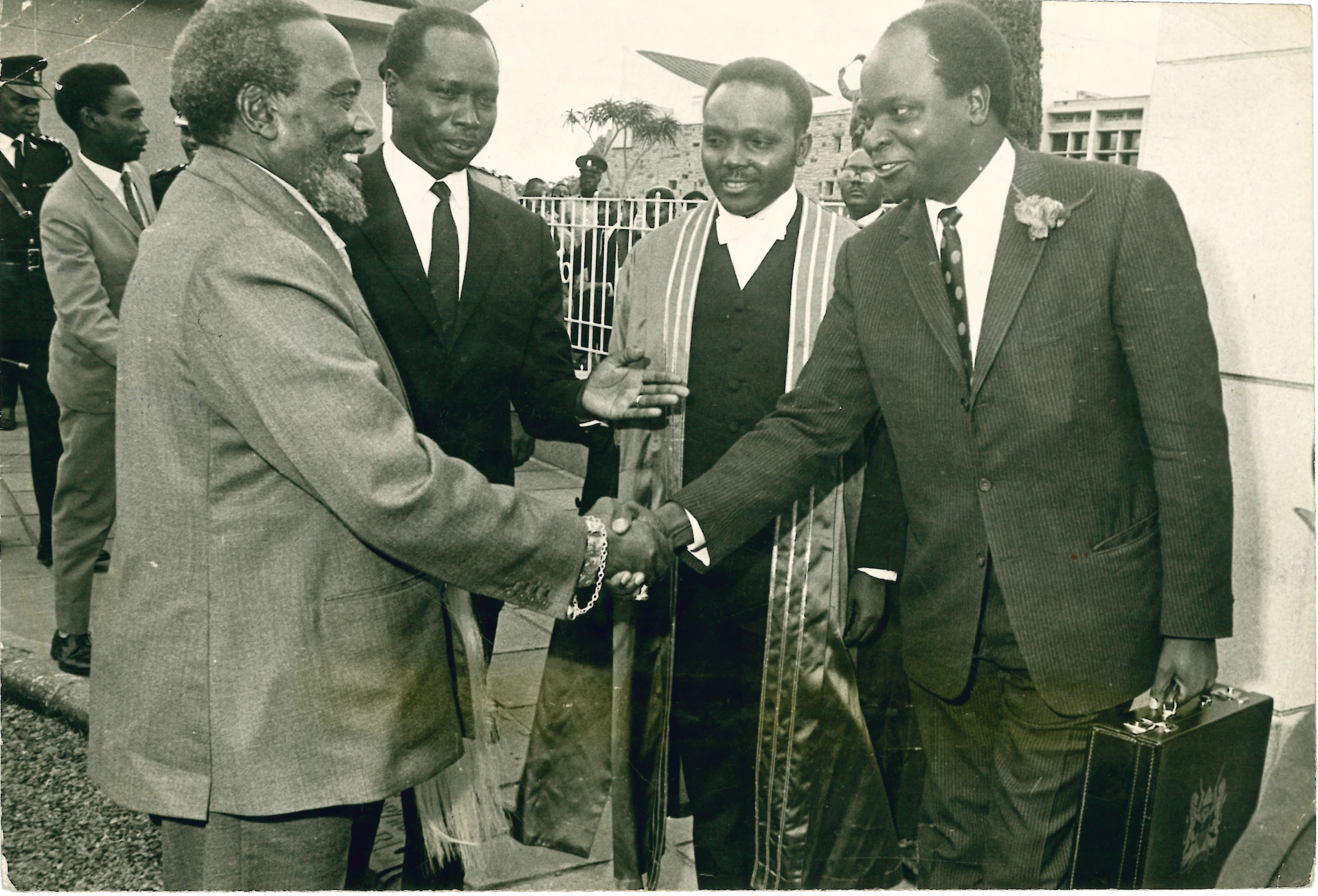 Black-and-white photo showing President Jomo Kenyatta shaking hand with Finance minister Mwai Kibaki wearing a dark suit, white tie and patterned tie, and holding the budget briefcase with his left hand. At the background, between the two, are Vice President Daniel arap Moi and the first African Clerck of the National Assembly Leonard James Ngugi in his official parliamentary attire.