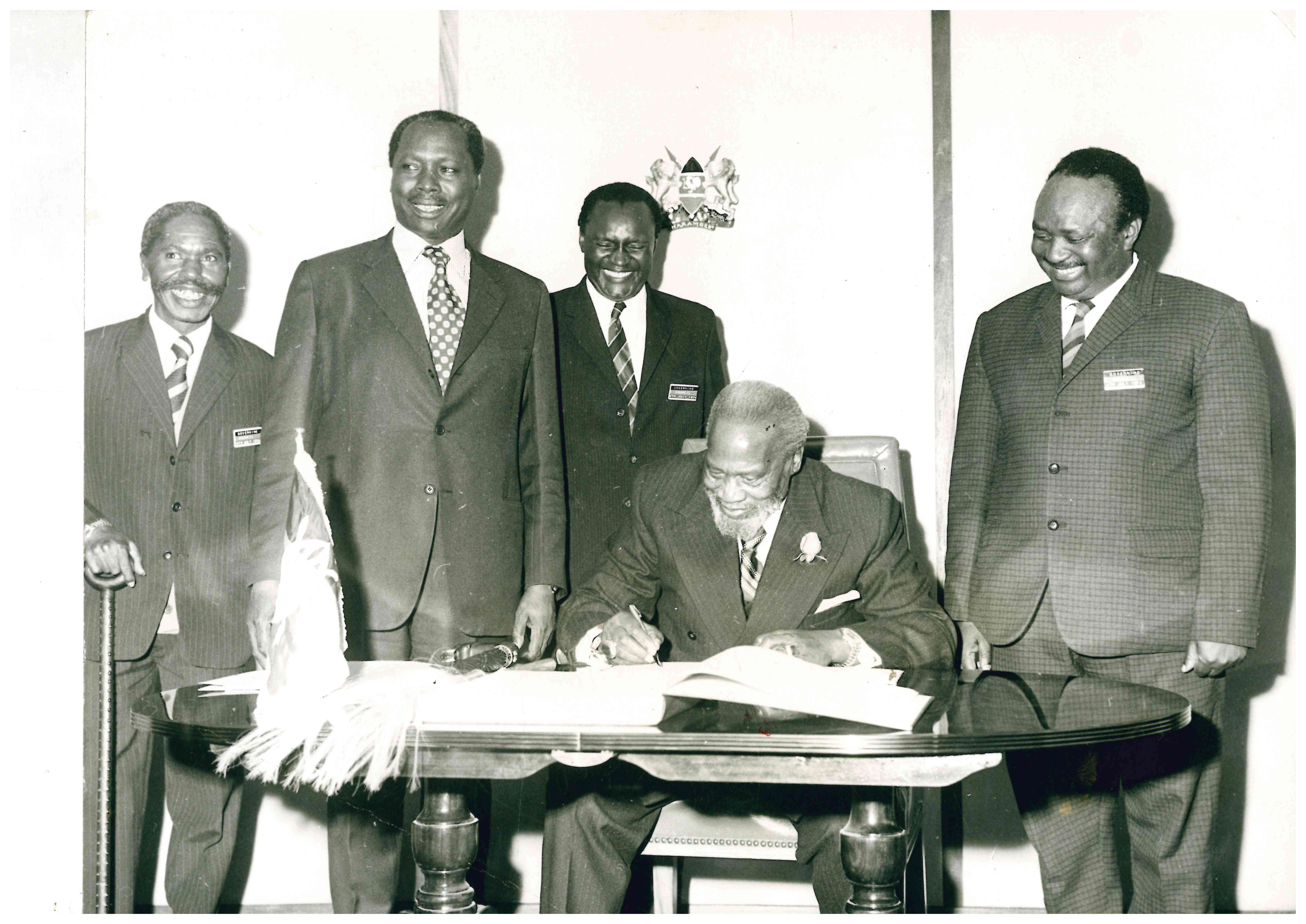 Black-and-white photo showing President Jomo Kenyatta signing an official document as his Vice Daniel Moi and other State officials stand round him all smiles. Kenyatta wears a dark suit, striped tie and flower lapel pin, while the officials are dressed in formal suits some with conference name badges. Kenyatta's ceremonial fly whisk rests on the table infront of him.