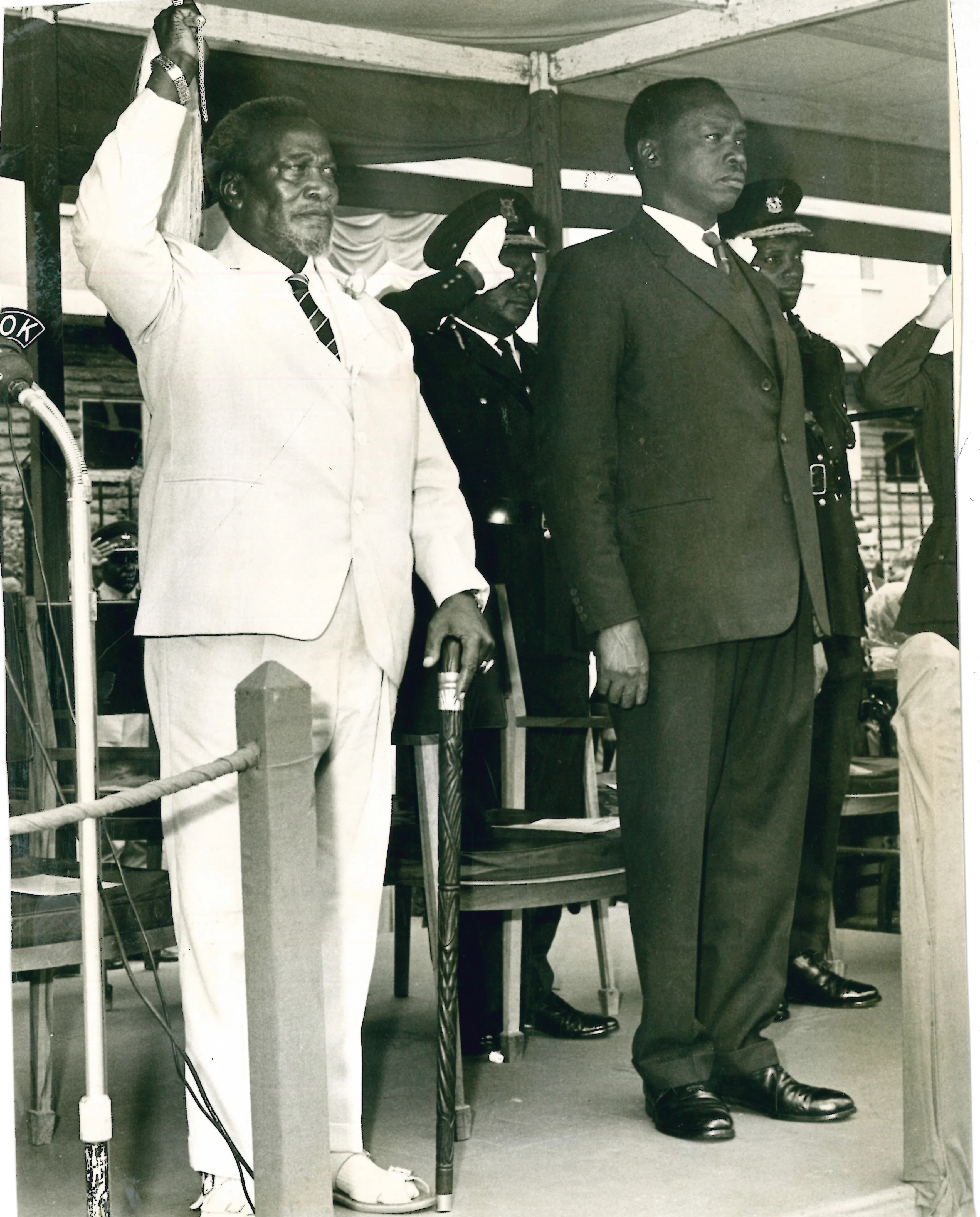 Black-and-white photo of President Jomo Kenyatta in a light-coloured suit, raising his ceremonial fly whisk during the 1967 Madaraka Day celebration on June 1. To his left is Vice President Daniel arap Moi in black suit and standing attention. In the background are saluting officers.
